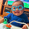 baby, child, infant, smiling, glasses, blue_glasses, portrait, indoor, playroom, toy, activity_center, high_chair, blue_shirt, television, sports, furniture, happy, sitting, colorful, family_home