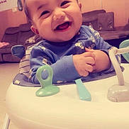 Adrian is registered to the contest to win money with this photo: baby, child, smiling, happy, portrait, indoor, living_room, activity_center, jumper, toy, infant, face, hands, blue_clothing, floral_pattern, couch, furniture, home, sitting, family_photo