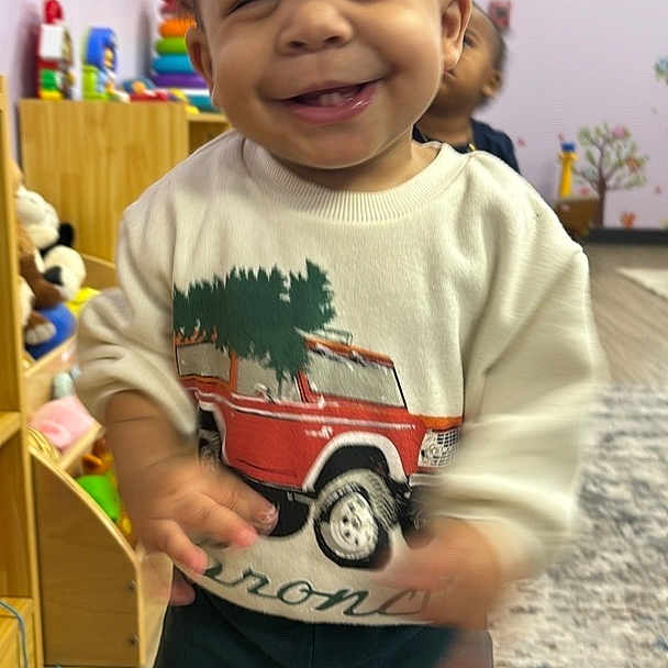 Syair is registered to the contest to win money with this photo: toddler, child, smiling, indoor, playroom, toys, clothing, sweater, pants, car_print, happy, motion_blur, person, floor, rug, wood_floor, furniture, background, young_child, cute