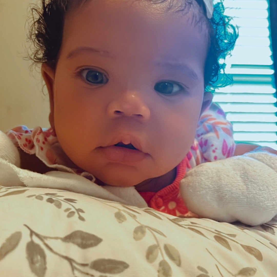 Jaci is registered to the contest to win money with this photo: adorable, baby, child, close_up, curiosity, curly_hair, cute, expression, eyes, face, floral_pattern, hair_clip, indoor, infant, natural_light, person, pillow, resting, soft_lighting, young