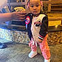 toddler, child, indoor, person, standing, holding_hand, jacket, pants, sneakers, hair_accessories, birthday_badge, carpet, stone_wall, adult_hand, fashion, cute, young_child, portrait, celebration, smiling
