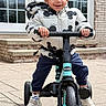 activity, boy, brick_wall, casual_clothing, child, daytime, door, happy, jacket, mickey_mouse, outdoor, pavement, person, play, shoes, smile, toy, tricycle, window, young