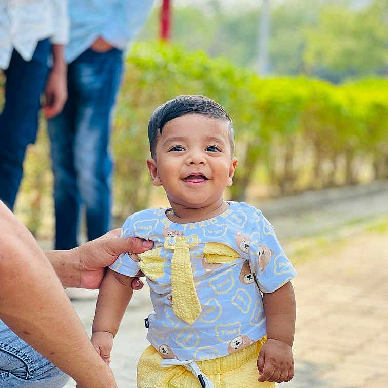 Manaf is registered to the contest to win money with this photo: adult_hand, blue_clothing, casual, child, cute, greenery, happy, hedge, outdoor, pavement, person, shorts, smiling, standing, sunlight, tie_print, toddler, tshirt, yellow_clothing, young_child