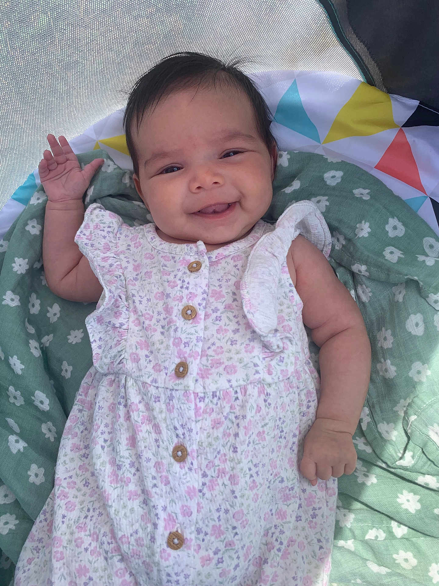 Veronica joined the competition — help win amazing prizes! baby, smiling, floral_dress, blanket, green, pattern, hand, face, cheerful, lying_down, infant, cute, natural_light, mesh_canopy, wooden_buttons, small_hand, happy, young_child, portrait, indoors