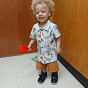 Kayden is registered to the contest to win money with this photo: toddler, child, indoor, smiling, curly_hair, toy, shirt, shorts, shoes, floor, door, wood_paneling, book, happy, play, person, standing, casual_clothing, red_toy, light