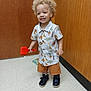 toddler, child, indoor, smiling, curly_hair, toy, shirt, shorts, shoes, floor, door, wood_paneling, book, happy, play, person, standing, casual_clothing, red_toy, light