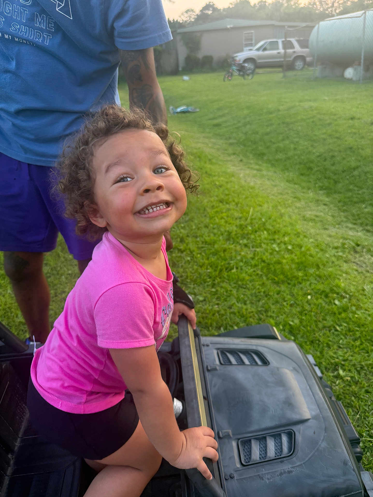 Lillyana joined the competition — help win amazing prizes! adult, backyard, black_shorts, casual_clothing, child, curly_hair, daylight, fun, grass, greenery, happy, outdoor, person, pink_shirt, play, smile, sunlight, toddler, toy_car, vehicle