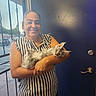 Camila is registered to the contest to win money with this photo: woman, cat, indoor, striped_clothing, smiling, holding, door, window, metal_grid, feline, pet, casual, portrait, happy, light, wall, home, hair_tied, sleeveless, domestic