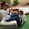 woman, dog, outdoor, wicker_chair, cushion, artificial_grass, garden, plant, bush, firepit, casual_clothing, glasses, sitting, pet, red_collar, leisure, daytime, side_profile, human, animal