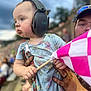 Elora joined the competition — help win amazing prizes! adult_hand, blue_shirt, butterfly_print, child, crowd, ear_protection, event, expression, flag, focus, headwear, holding, outdoor, patterned_clothing, person, sky, spectator, stadium, support, toddler