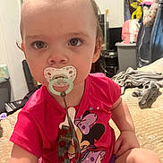 Elora is registered to the contest to win money with this photo: bed, blanket, child, clothing, curious, expression, eyes, face, furniture, hair, hand, indoor, minnie_mouse, pacifier, person, red_shirt, room, toddler, toy, young_child