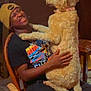 man, dog, beanie, chair, indoor, casual, smiling, fluffy_dog, wooden_chair, black_tshirt, happy, pet, playing, heart_emojis, collar, holding, lap, pants, furniture, relaxed