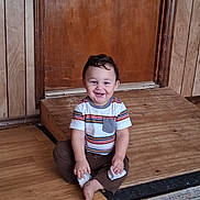 George is registered to the contest to win money with this photo: toddler, child, smiling, sitting, striped_shirt, brown_pants, wooden_floor, wooden_door, indoor, barefoot, happy, cute, portrait, person, young_child, flooring, clothing, sock, playful, home