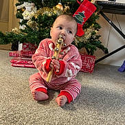 Iris is registered to the contest to win money with this photo: baby, child, christmas_tree, holiday, pajamas, carpet, present, gift, jingle_bells, wooden_toy, decorations, festive, indoors, floor, red_clothing, striped_pants, holiday_season, cute, winter_wear, sitting