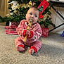 baby, child, christmas_tree, holiday, pajamas, carpet, present, gift, jingle_bells, wooden_toy, decorations, festive, indoors, floor, red_clothing, striped_pants, holiday_season, cute, winter_wear, sitting