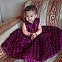 toddler, child, purple_dress, sofa, patterned_sofa, henna, clothing, indoor, sitting, curious, small_child, furniture, decor, floor, wooden_floor, cute, portrait, person, young_child, smiling