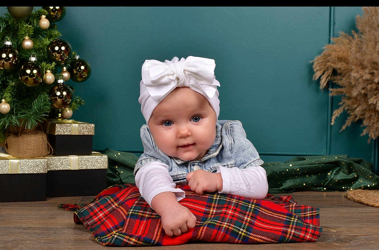 Savanna participe au concours pour gagner de l'argent avec cette photo : baby, infant, child, white_bow, denim_jacket, plaid_blanket, christmas_tree, gold_baubles, presents, holiday_decor, teal_wall, floor, wooden_floor, posing, cute, portrait, smiling, indoor, festive, cozy