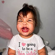 Emily is registered to the contest to win money with this photo: toddler, child, smiling, happy, shirt, text, indoor, portrait, baby, necklace, cute, funny_expression, hair, pink, white, wall_decor, sitting, person, clothing, celebration