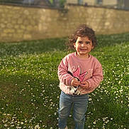 Melissa participe au concours pour gagner de l'argent avec cette photo : building, child, curly_hair, cute, daylight, flowers, grass, greenery, happy, jeans, nature, outdoor, person, shoes, smiling, standing, sunlight, sweater, wall, yard
