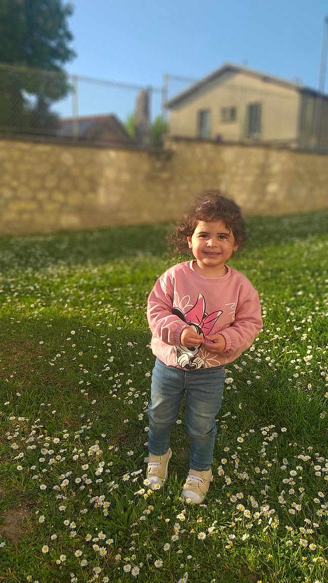 Melissa participe au concours pour gagner de l'argent avec cette photo : building, child, curly_hair, cute, daylight, flowers, grass, greenery, happy, jeans, nature, outdoor, person, shoes, smiling, standing, sunlight, sweater, wall, yard