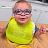 Jackson is registered to the contest to win money with this photo: toddler, child, glasses, green_bib, smiling, thumbs_up, eating, food, plate, table, kitchen, indoor, person, baby, happy, cute, wearing, face, sitting, mealtime