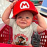 baby, child, shopping_cart, red_hat, mario_hat, onesie, toy_store, aisle, smiling, cute, face, person, clothing, infant, baby_clothes, shopping, red, cart, indoor, cute_expression