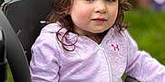 Brynnlee joined the competition — help win amazing prizes! child, curly_hair, purple_hoodie, high_chair, outdoor, green_background, face, person, young_child, sitting, hand, table, casual_clothing, daylight, portrait, expression, looking_away, soft_lighting, close_up, cute