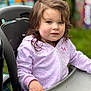 child, curly_hair, purple_hoodie, high_chair, outdoor, green_background, face, person, young_child, sitting, hand, table, casual_clothing, daylight, portrait, expression, looking_away, soft_lighting, close_up, cute