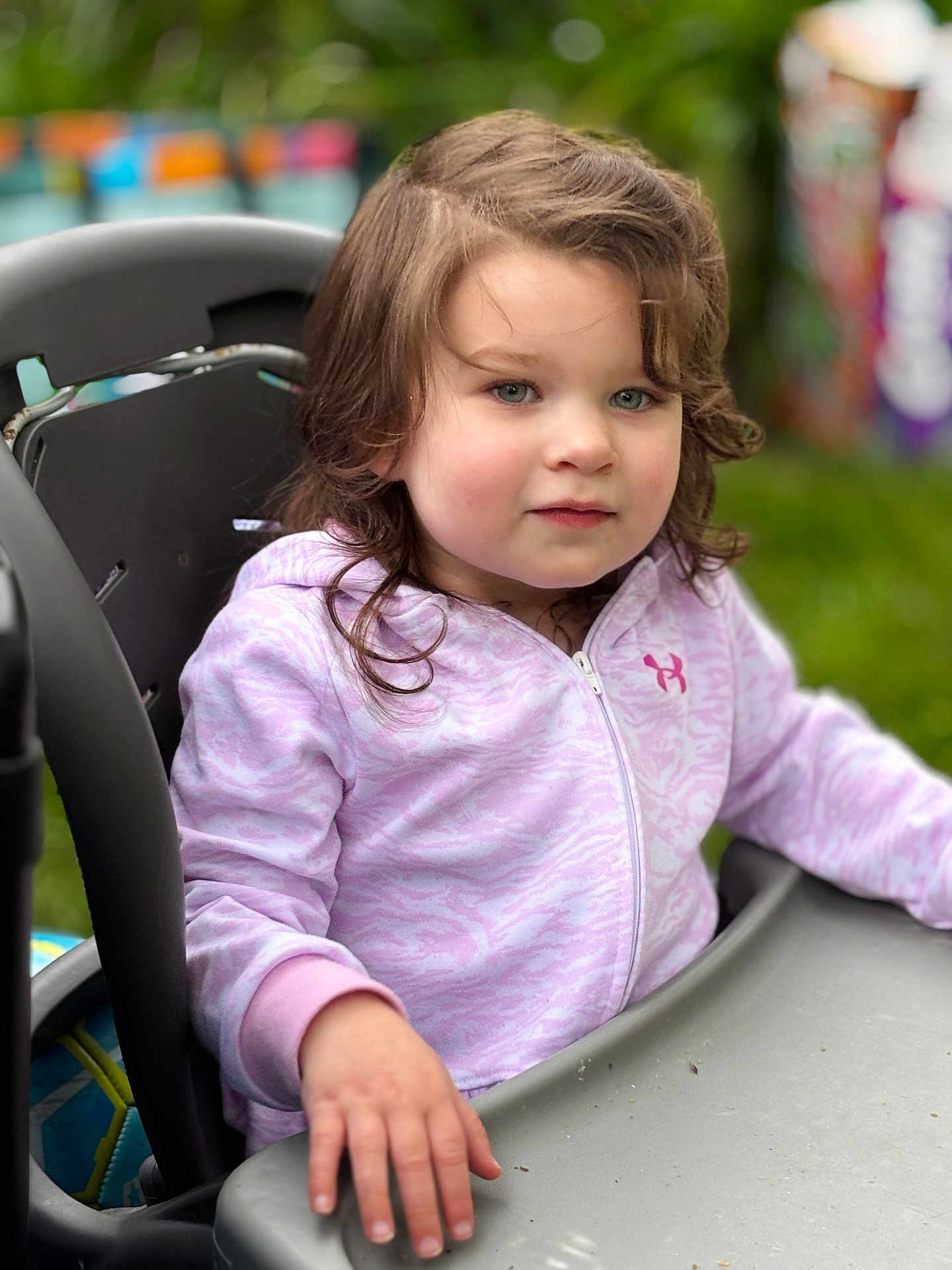 Brynnlee joined the competition — help win amazing prizes! child, curly_hair, purple_hoodie, high_chair, outdoor, green_background, face, person, young_child, sitting, hand, table, casual_clothing, daylight, portrait, expression, looking_away, soft_lighting, close_up, cute