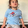 Brynnlee joined the competition — help win amazing prizes! child, toddler, sitting, blue_tshirt, shorts, patterned_clothing, hands, wooden_surface, plain_background, curly_hair, looking_up, casual_clothing, indoor, young_child, portrait, innocent_expression, candid, light_skin, daylight, clothing_design