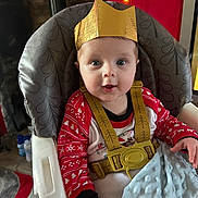 Kaleb is registered to the contest to win money with this photo: baby, blanket, blue_blanket, carpet, child, crown, decorations, face, festive, happy, high_chair, holiday_theme, indoor, living_room, patterned_clothing, person, photography, red_clothing, seat_belt, smiling