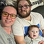 family, selfie, woman, man, baby, glasses, beard, couch, pillow, text, indoor, smile, happy, casual_clothing, striped_clothing, people, portrait, togetherness, home, cozy