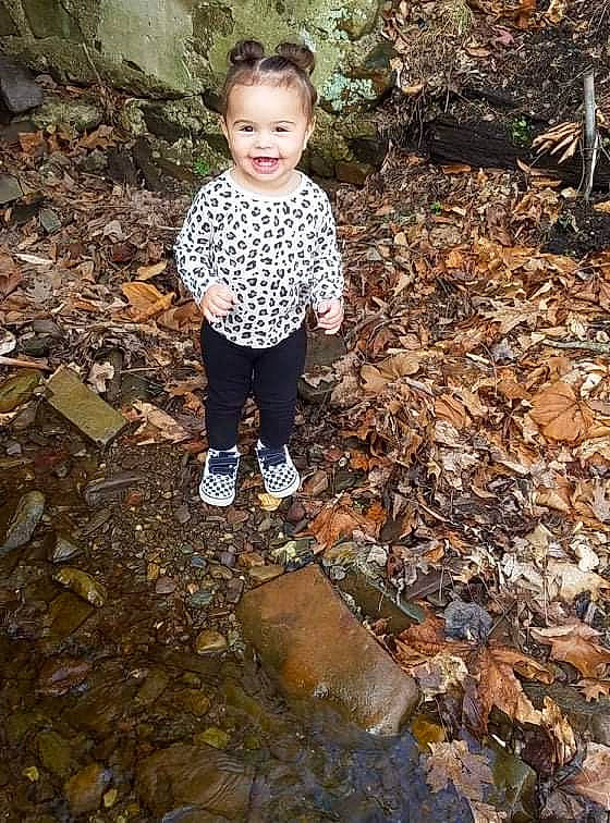 Mariana is registered to the contest to win money with this photo: asphalt, child, cobblestone, deciduous, forest, grass, joy, landscape, leaf, leg, people_in_nature, person, road_surface, rock, smile, soil, standing, toddler, tree, water