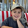 child, boy, smile, baseball_cap, hat, face, portrait, hand_on_face, menu, table, striped_chair, wooden_fence, bokeh, eyes, teeth, casual_shirt, happy, sitting, restaurant, young_child