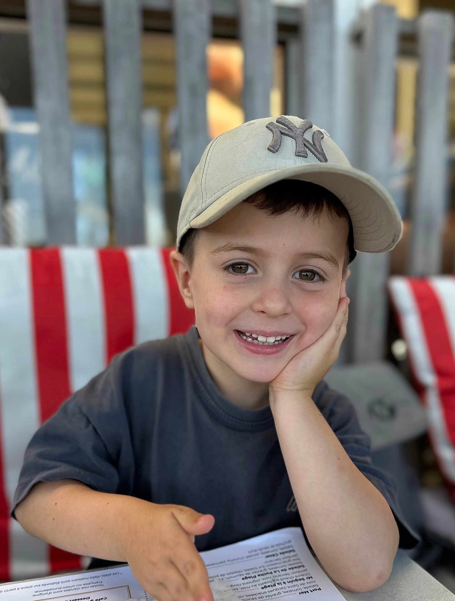 Naël a rejoint le concours — aidez-le/la à gagner de superbes lots ! child, boy, smile, baseball_cap, hat, face, portrait, hand_on_face, menu, table, striped_chair, wooden_fence, bokeh, eyes, teeth, casual_shirt, happy, sitting, restaurant, young_child