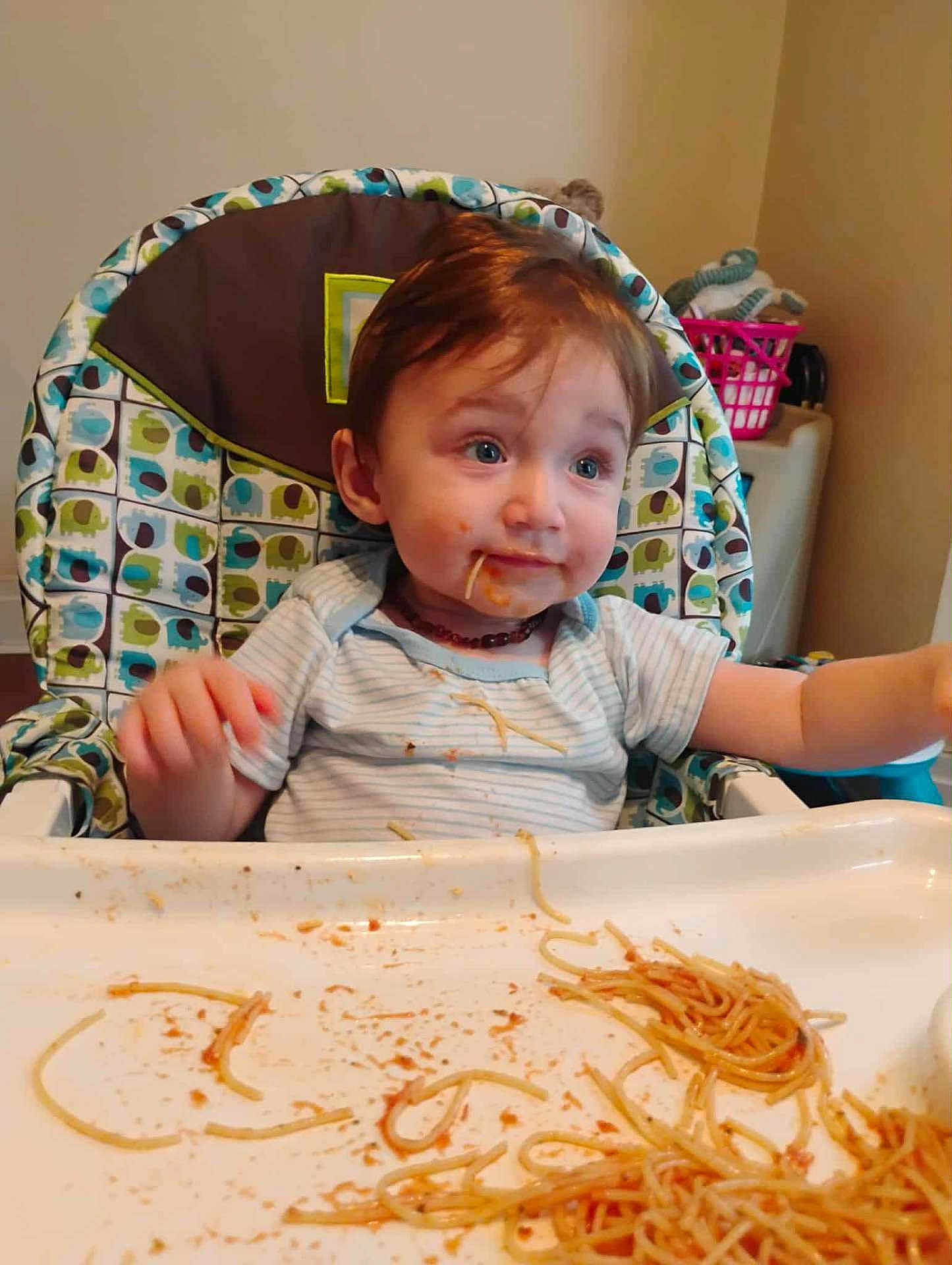 Otis is registered to the contest to win money with this photo: child, baby, high_chair, spaghetti, messy, food, meal, indoor, face, clothing, plate, tray, hair, blue_eyes, expression, sitting, cute, kitchen, dining, feeding