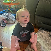 Truman is registered to the contest to win money with this photo: baby, child, sitting, floor, toy, handle, tshirt, rolling_stones, hair, pony_tail, living_room, carpet, furniture, curious, indoor, wooden_floor, clothing, person, hand, foot