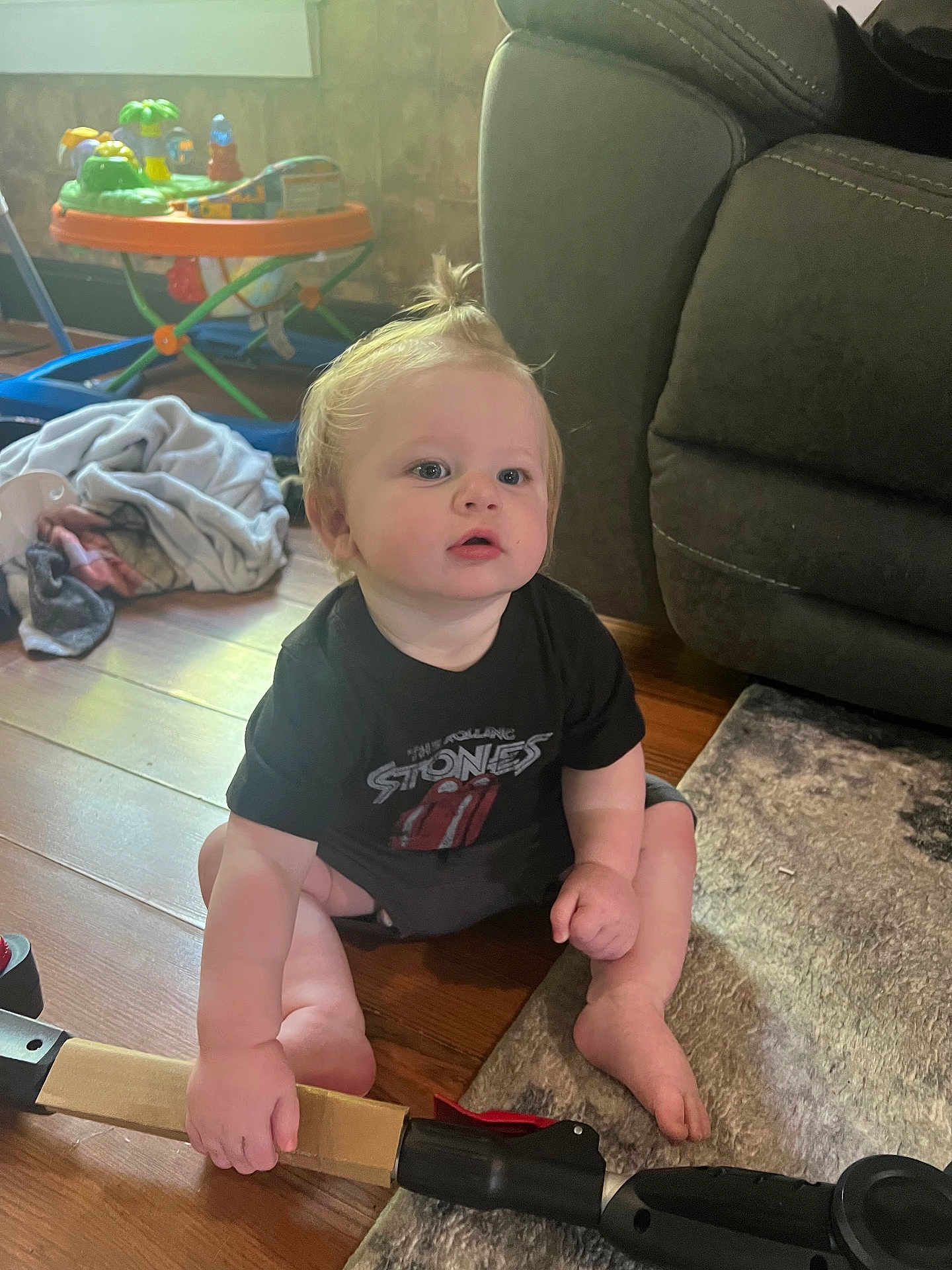 Truman is registered to the contest to win money with this photo: baby, child, sitting, floor, toy, handle, tshirt, rolling_stones, hair, pony_tail, living_room, carpet, furniture, curious, indoor, wooden_floor, clothing, person, hand, foot