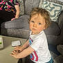 child, toddler, curly_hair, white_tshirt, blue_pants, table, sofa, cushions, older_woman, glasses, living_room, carpet, indoor, person, casual_clothing, face, hands, expression, home, relaxing