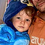 child, toddler, adult, hoodie, blue_clothing, orange_clothing, face, portrait, indoor, closeup, cuddle, casual, expression, warmth, curly_hair, window, light, person, smile, cozy