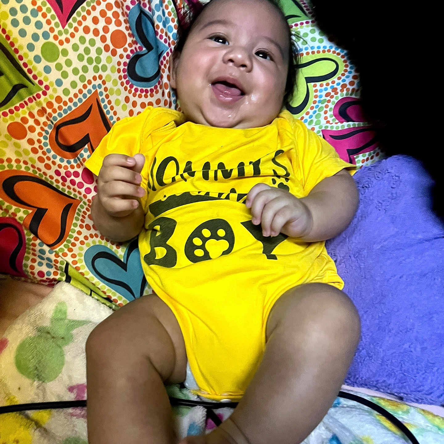 Kingston is registered to the contest to win money with this photo: baby, infant, child, smiling, happy, portrait, indoors, bed, bedding, colorful, yellow_outfit, onesie, lying_down, sitting, face, hands, feet, pillow, heart_pattern, playful