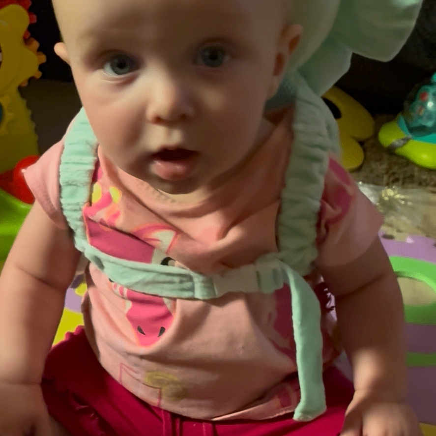 Zoey joined the competition — help win amazing prizes! baby, baby_toys, casual_clothing, child, colorful, curious, cushion, floor, harness, indoor, infant, open_mouth, person, pink_shirt, play_mat, red_shorts, sitting, toy, wide_eyes, young_child