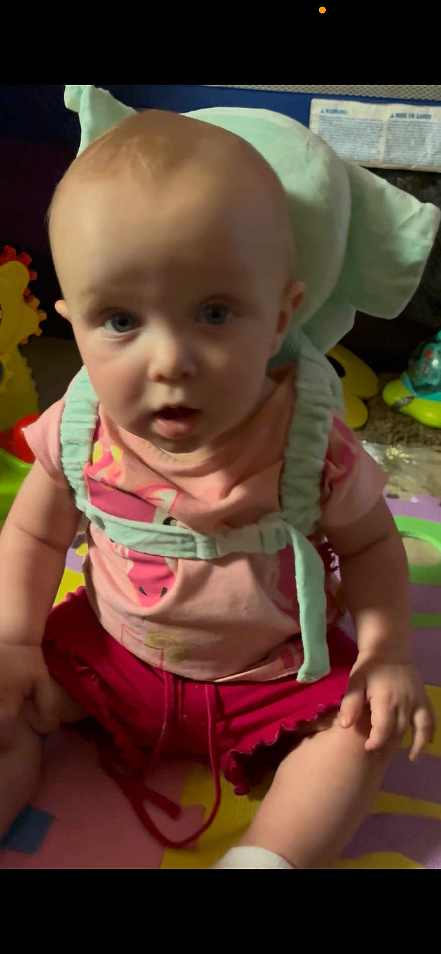 Zoey joined the competition — help win amazing prizes! baby, child, pink_shirt, red_shorts, harness, play_mat, toy, cushion, indoor, person, infant, sitting, curious, wide_eyes, open_mouth, colorful, floor, young_child, casual_clothing, baby_toys