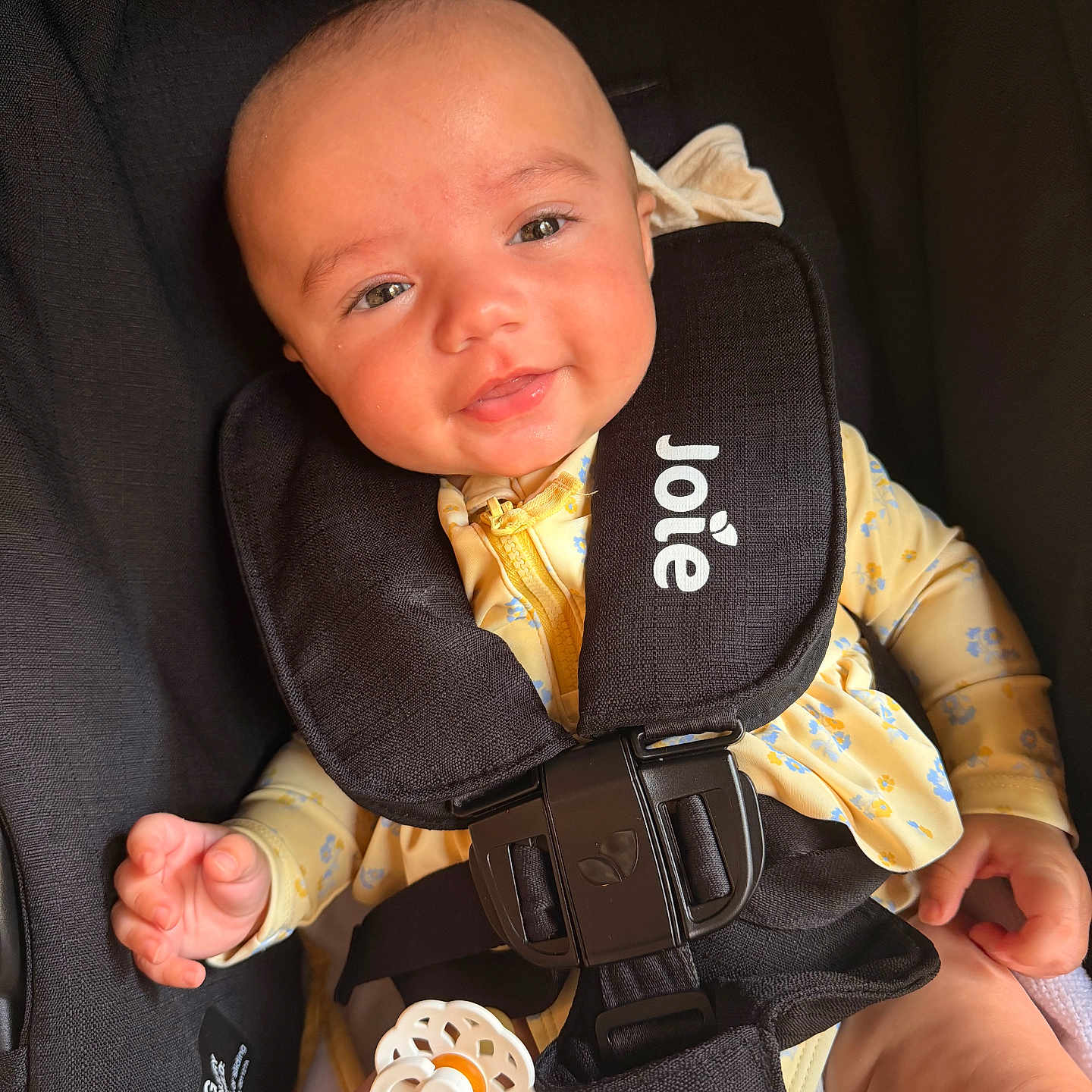 Emerie is registered to the contest to win money with this photo: baby, infant, car_seat, harness, pacifier, yellow_clothing, smile, face, child, safety, seatbelt, cute, young_child, person, apparel, comfort, indoor, closeup, head, hand