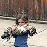 Ellie is registered to the contest to win money with this photo: child, cat, tabby_cat, outdoor, concrete, wooden_fence, casual_clothing, blue_shirt, jeans, holding, pet, animal, walking, curious, calm, young, person, nature, daytime, companion