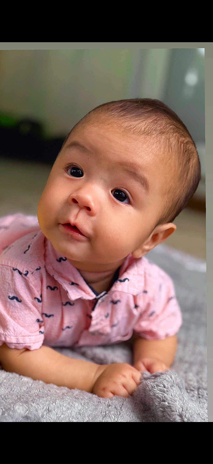 Mateo is registered to the contest to win money with this photo: baby, infant, child, face, eyes, pink_shirt, mustache_pattern, blanket, soft_texture, cute, portrait, indoor, skin, expression, hands, lying_down, young, head, curious, adorable