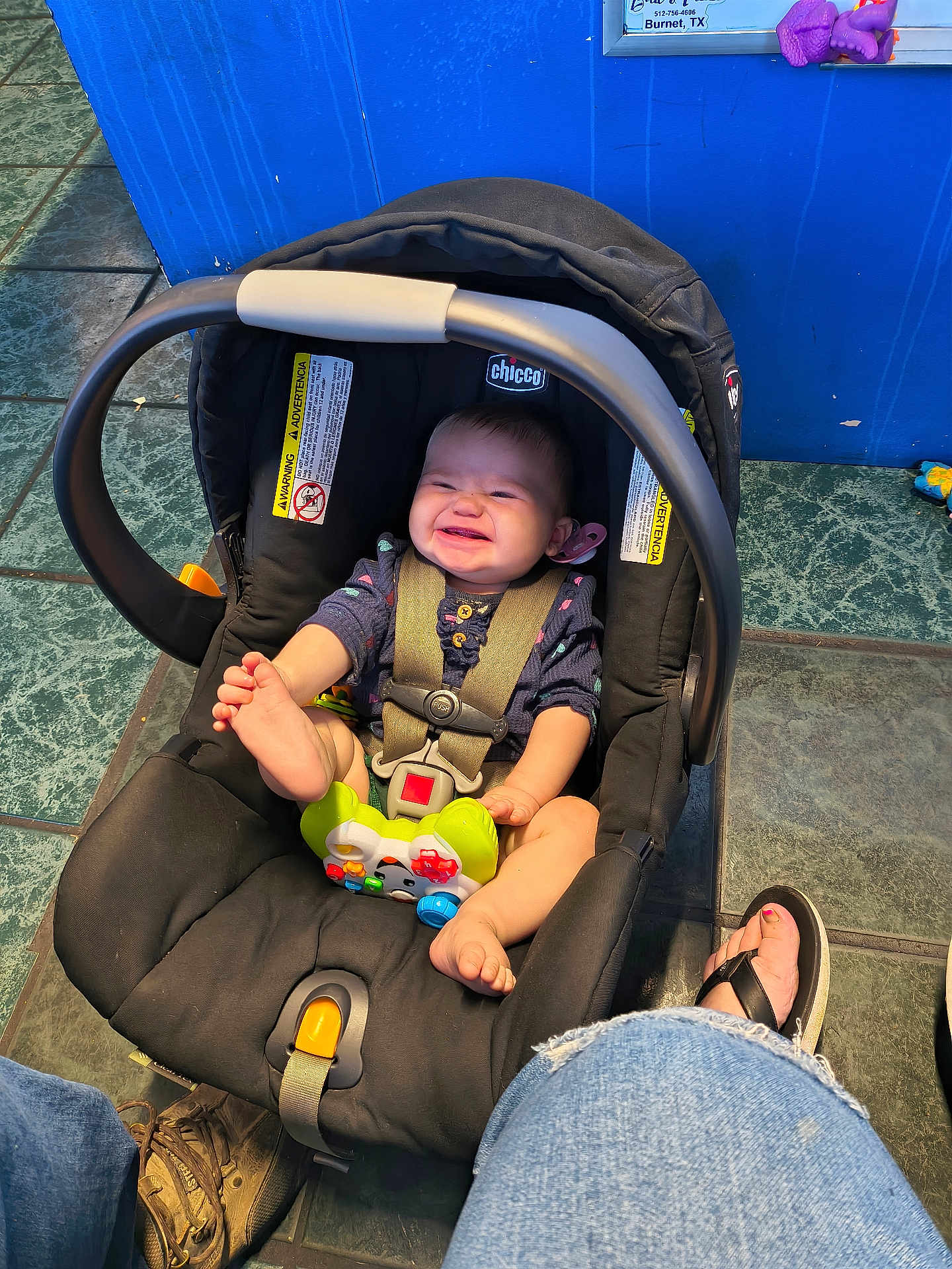 Olivia Grace Cowan is registered to the contest to win money with this photo: baby, car_seat, smiling, foot, toy, indoor, floor_tile, blue_wall, person, jeans, sandals, grin, straps, plastic_toy, child, casual, happy, seat, infant, playful