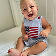 Weston joined the competition — help win amazing prizes! baby, child, smiling, sitting, indoor, bed, white, clothing, american_flag, cute, happy, portrait, infant, skin, feet, hand, face, blonde_hair, blue_eyes, room