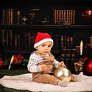 Nathan a rejoint le concours — aidez-le/la à gagner de superbes lots ! baby, child, toddler, santa_hat, christmas_ornament, holiday, festive, overalls, white_rug, candles, gifts, poinsettia, bookshelf, library, decorations, red, gold, indoors, portrait, smiling