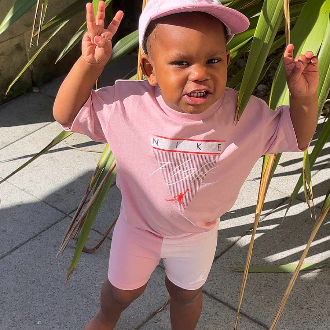 Loisse a rejoint le concours — aidez-le/la à gagner de superbes lots ! barefoot, cap, casual, child, concrete, gesture, greenery, happy, nike, outdoor, person, pink_clothing, plants, playful, shadow, sidewalk, smiling, sunlight, toddler, young_child